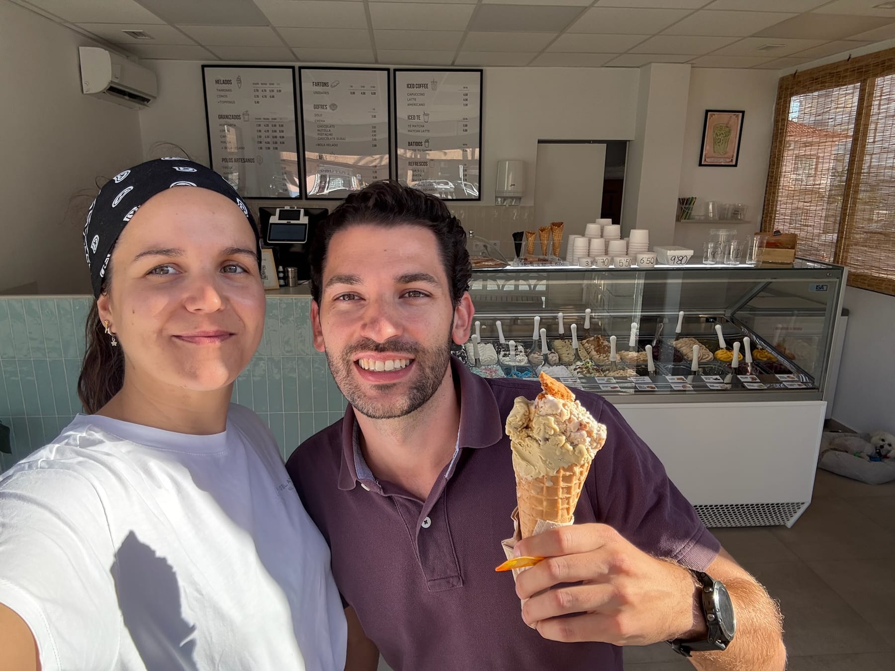 La propietaria y un cliente sonríen tras la vitrina; el cliente sostiene un cucurucho de helado