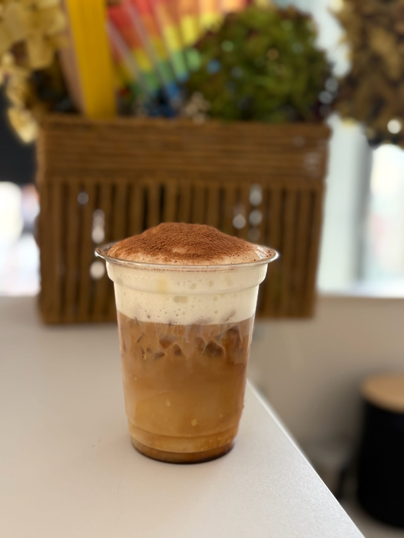 Iced coffee crowned with white foam dusted with cocoa, dried flowers in the background